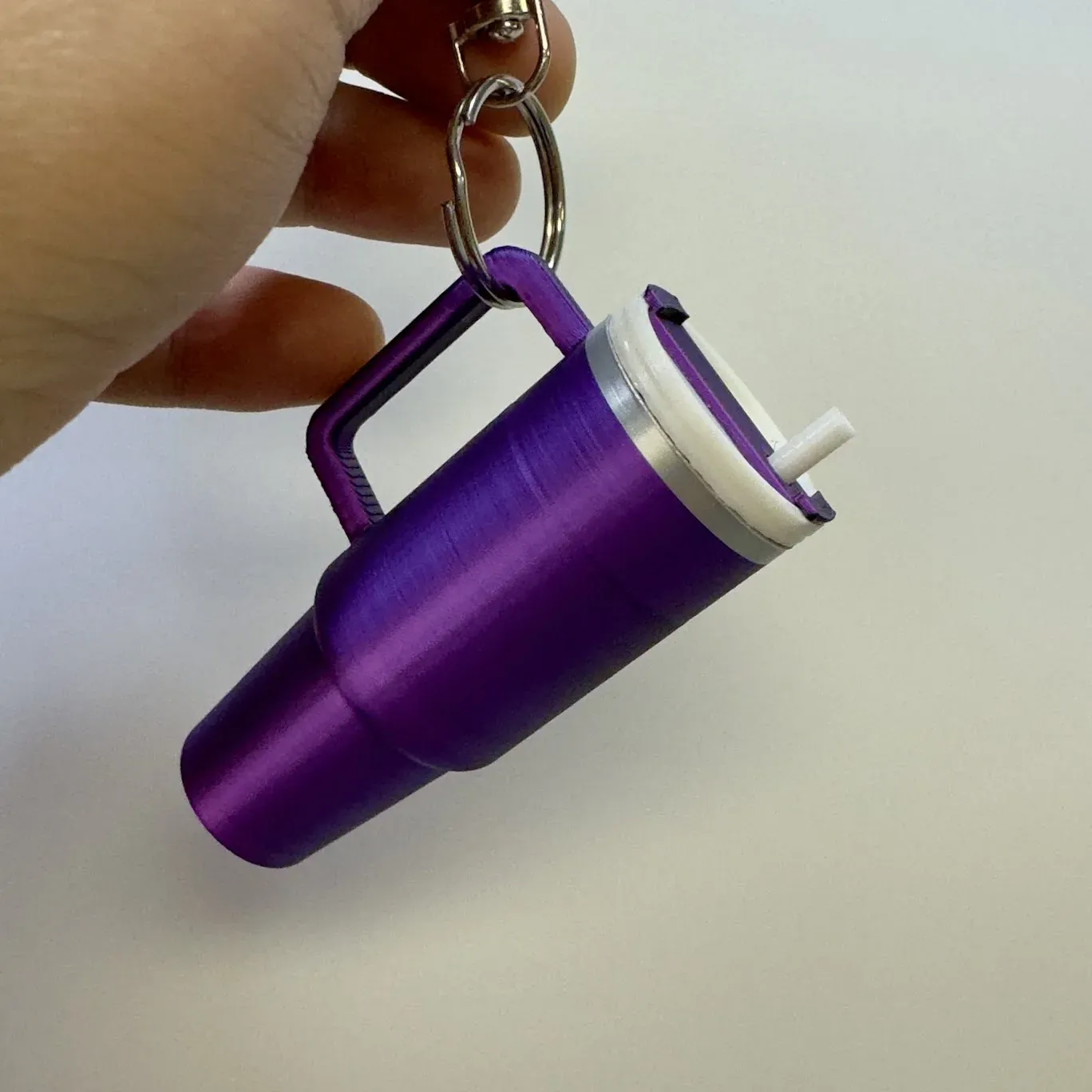 A hand holding a purple printed Stanley-style tumbler on a keyring, with a white lid and straw